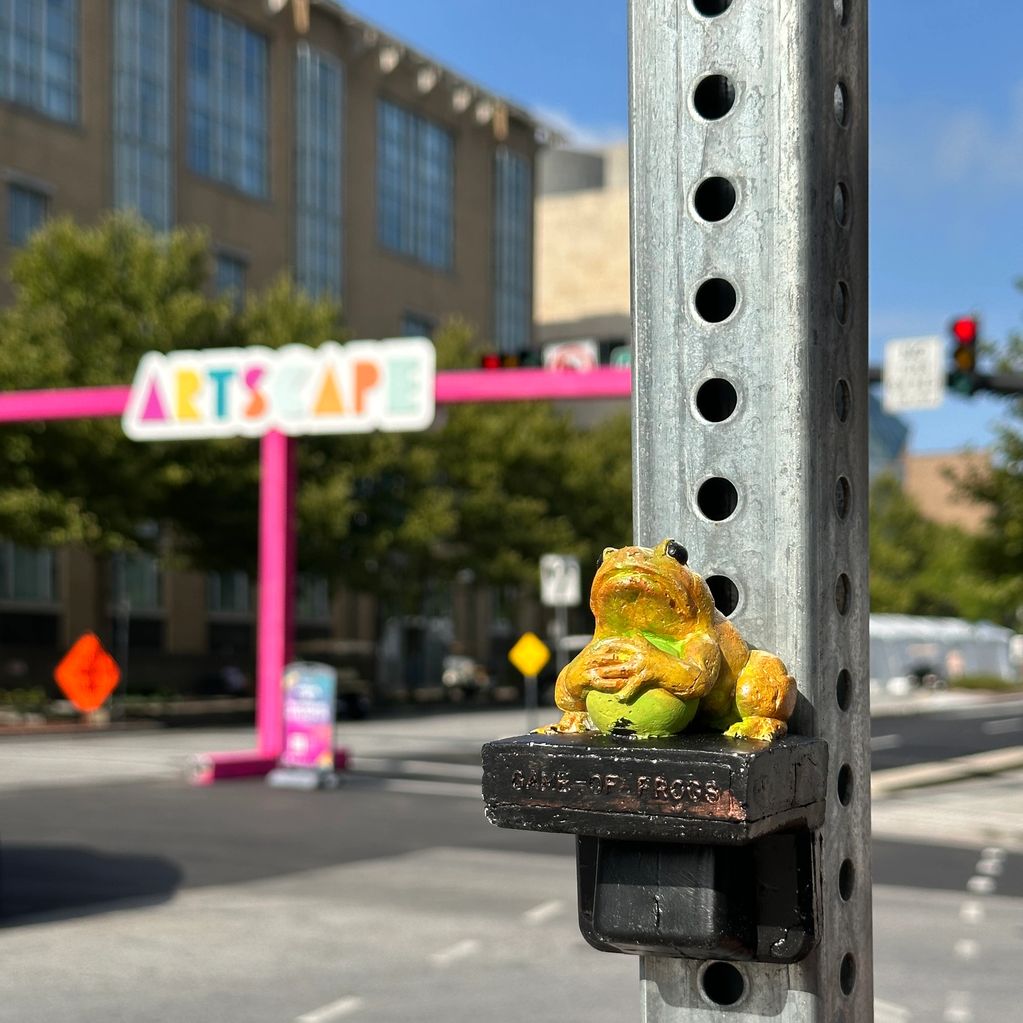 A small frog statue on a metal pole in an urban setting.
