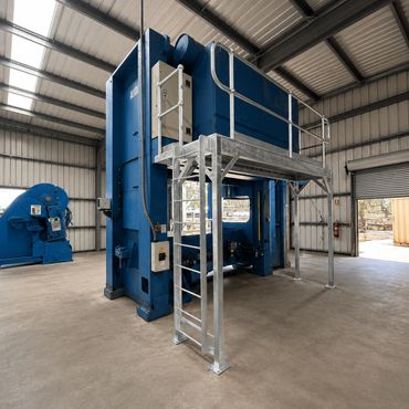 Large blue industrial machinery inside a warehouse with a metallic platform and ladder.