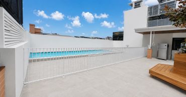 Modern outdoor pool area with safety fence and wooden seating under blue sky.