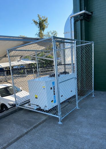 A white generator secured inside a chain-link fenced enclosure outdoors.
