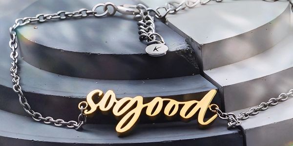 Silver chain bracelet with a gold 'so good' pendant on a black and white textured surface.