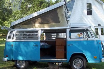 Blue vintage camper van with raised pop-up roof parked outside a white house.