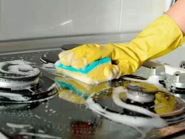 Hand wearing yellow glove scrubs a soapy stovetop with a sponge.