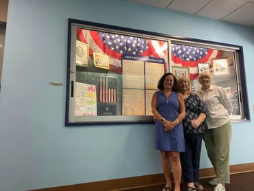 Three women standing in front of a patriotic display case with American flags and documents.