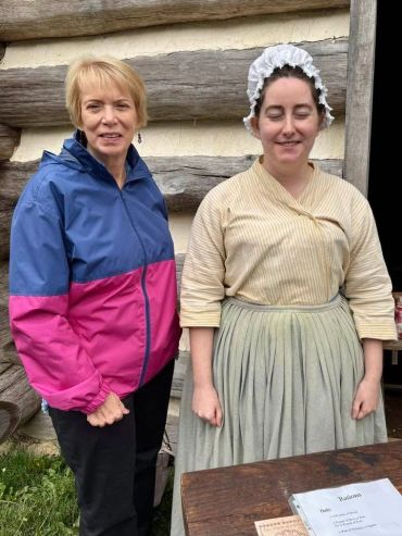 Two women standing outside a rustic wooden building, one in modern clothes, the other in historical attire.