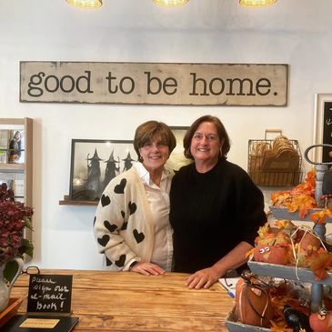 Two women smiling behind a wooden counter under a 'good to be home' sign.