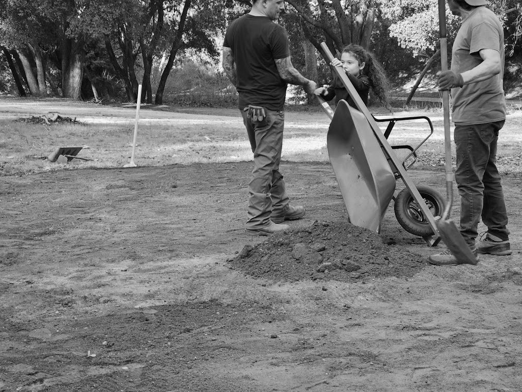 Child and adults working with soil and tools during Indigenous land stewardship in a forest clearing