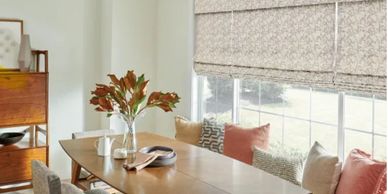 Bright dining room with wooden table, cushioned chairs, and large window with patterned blinds.
