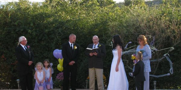 Outdoor wedding ceremony with bride, groom, officiant, and family members.