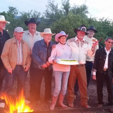 Group of people in cowboy hats gathered around a campfire, one holding a cake.