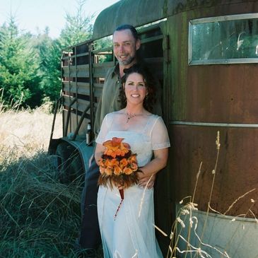 A couple in wedding attire posing outdoors by a rustic trailer.