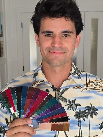 Smiling man holding a colorful fabric swatch fan, wearing a tropical shirt.