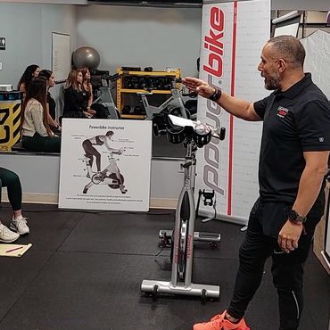 Instructor explains Powerbike indoor cycling to a small group in a gym.