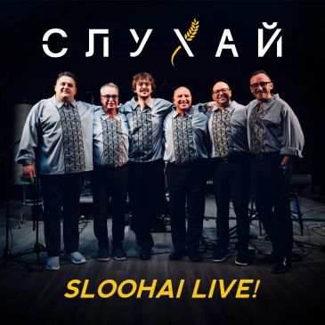 Six men in traditional embroidered shirts smiling together on stage.