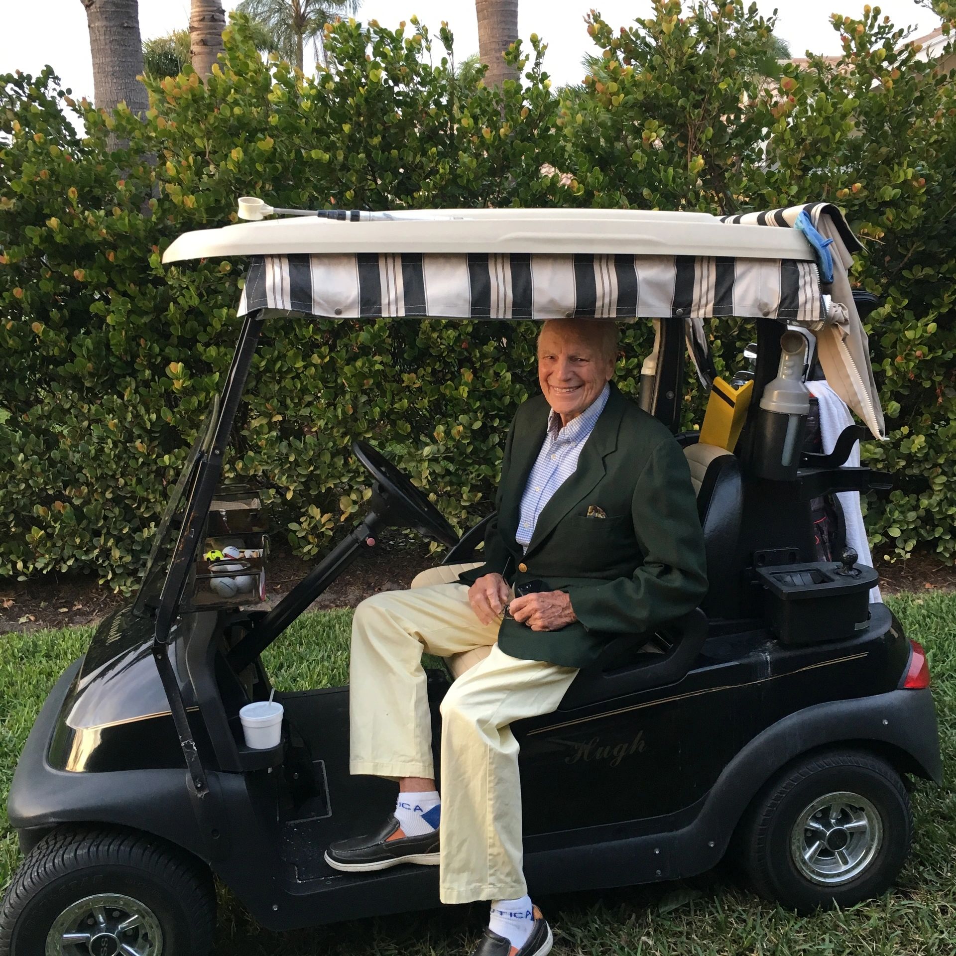 Elderly man smiling while sitting in a golf cart beside greenery.