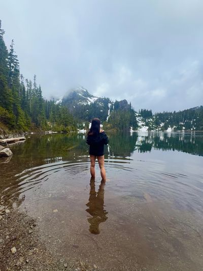 Person standing in a serene mountain lake surrounded by snow and trees.