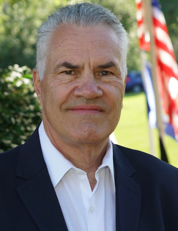 Smiling older man in white shirt and dark blazer with American flags in the background.