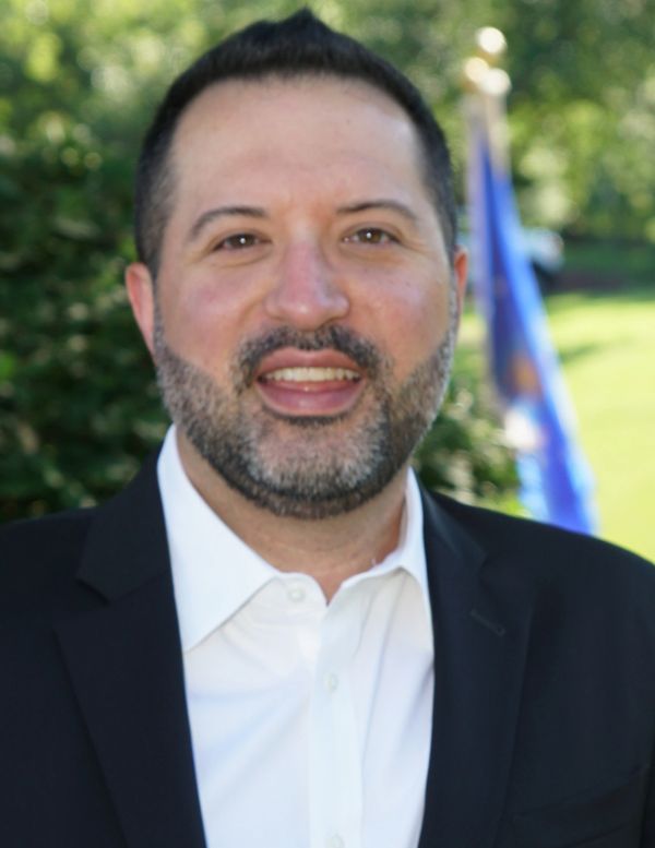 Man with beard wearing a black suit and white shirt outdoors.
