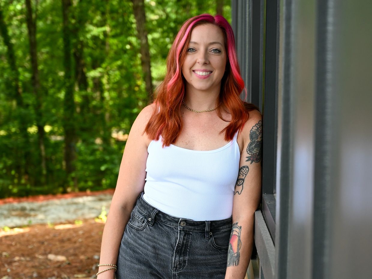 Young woman with pink and orange hair leans against a building smiling outdoors.