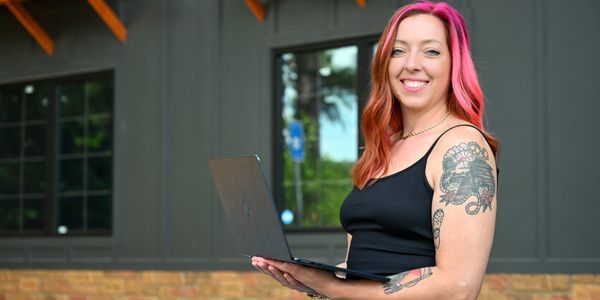 Smiling woman with pink hair holding a laptop outdoors.