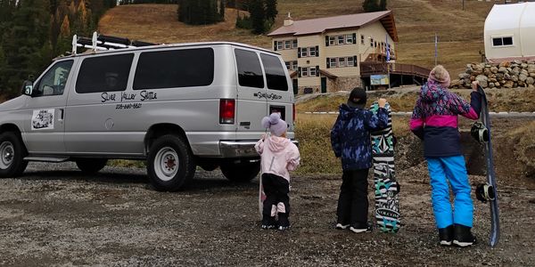 Three children with snowboards near a shuttle van in a mountainous area.
