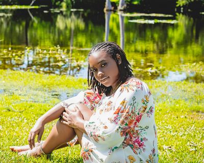 A woman in a floral dress sits barefoot by a pond, surrounded by greenery.