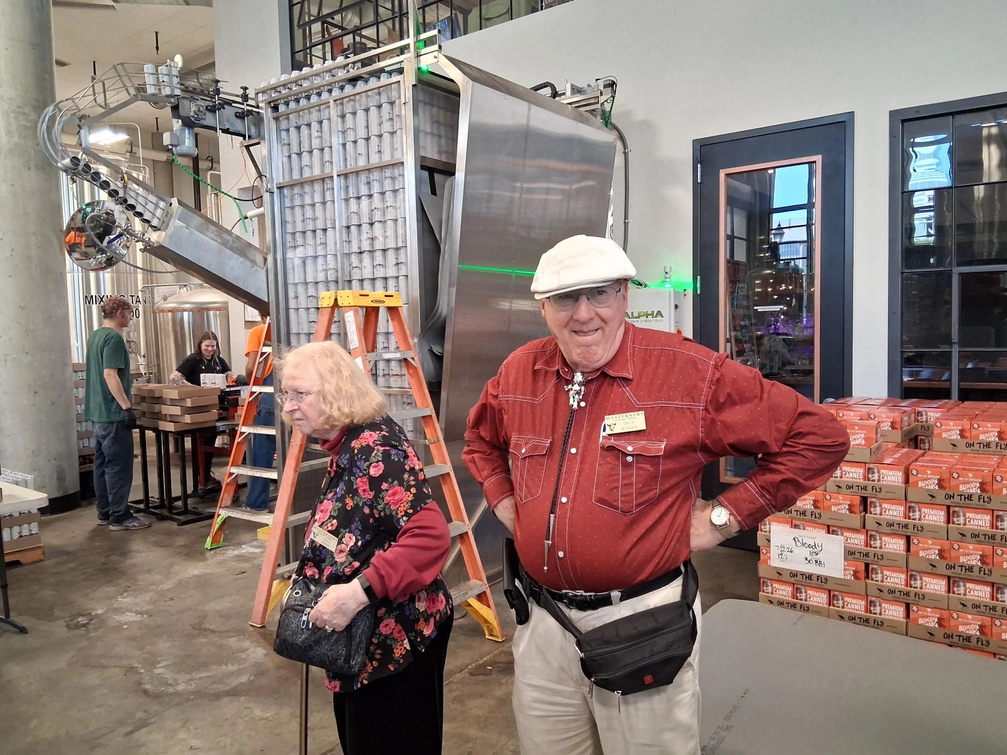Two elderly people in a factory setting with stacked canned goods.