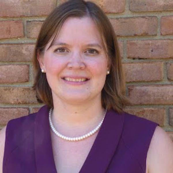 Smiling woman wearing a purple top and pearl necklace against a brick wall.