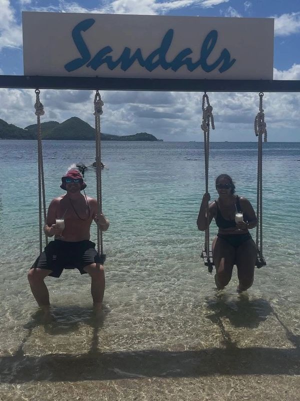 Two people on swings in the water under a 'Sandals' sign, enjoying drinks.
