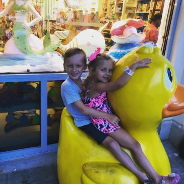 Two kids sitting on a large yellow rubber duck outside a store.