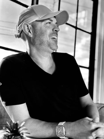 Smiling man wearing a cap and watch, sitting by a window.
