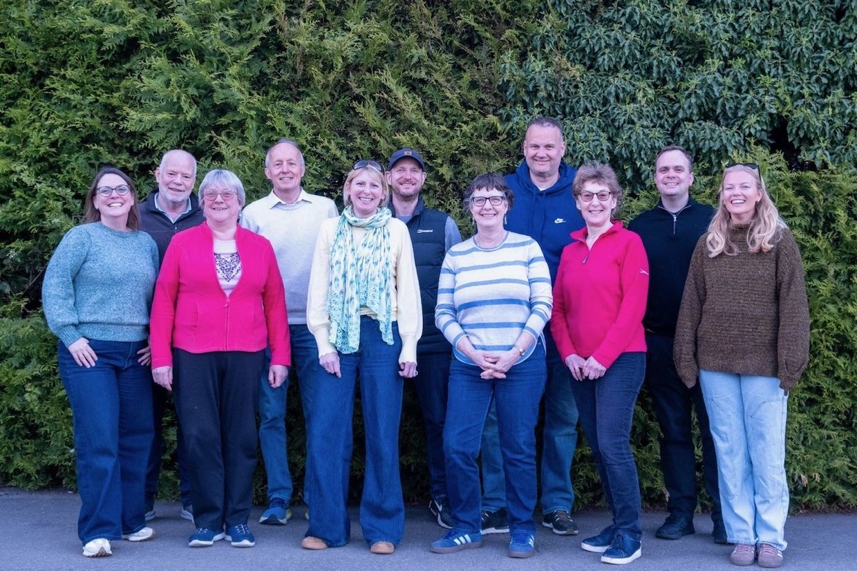 A group of eleven adults standing outdoors in front of dense green shrubbery, smiling at the camera.