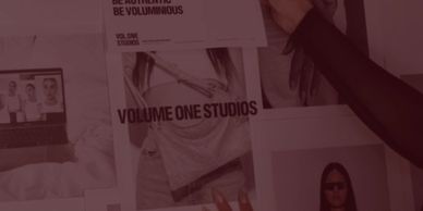 A hand holding a "VOLUME ONE STUDIOS" poster among fashion photos and a laptop.