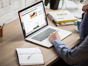 Person using a laptop on a wooden desk with a notebook and coffee.