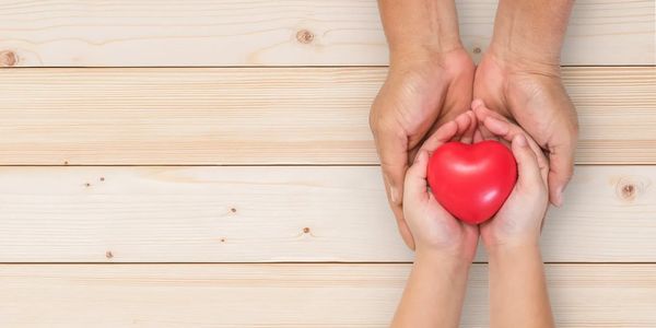 Adult hands holding child hands with a red heart on wooden table.