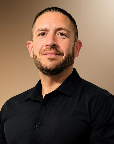 Confident man with a trimmed beard wearing a black shirt against a beige background.