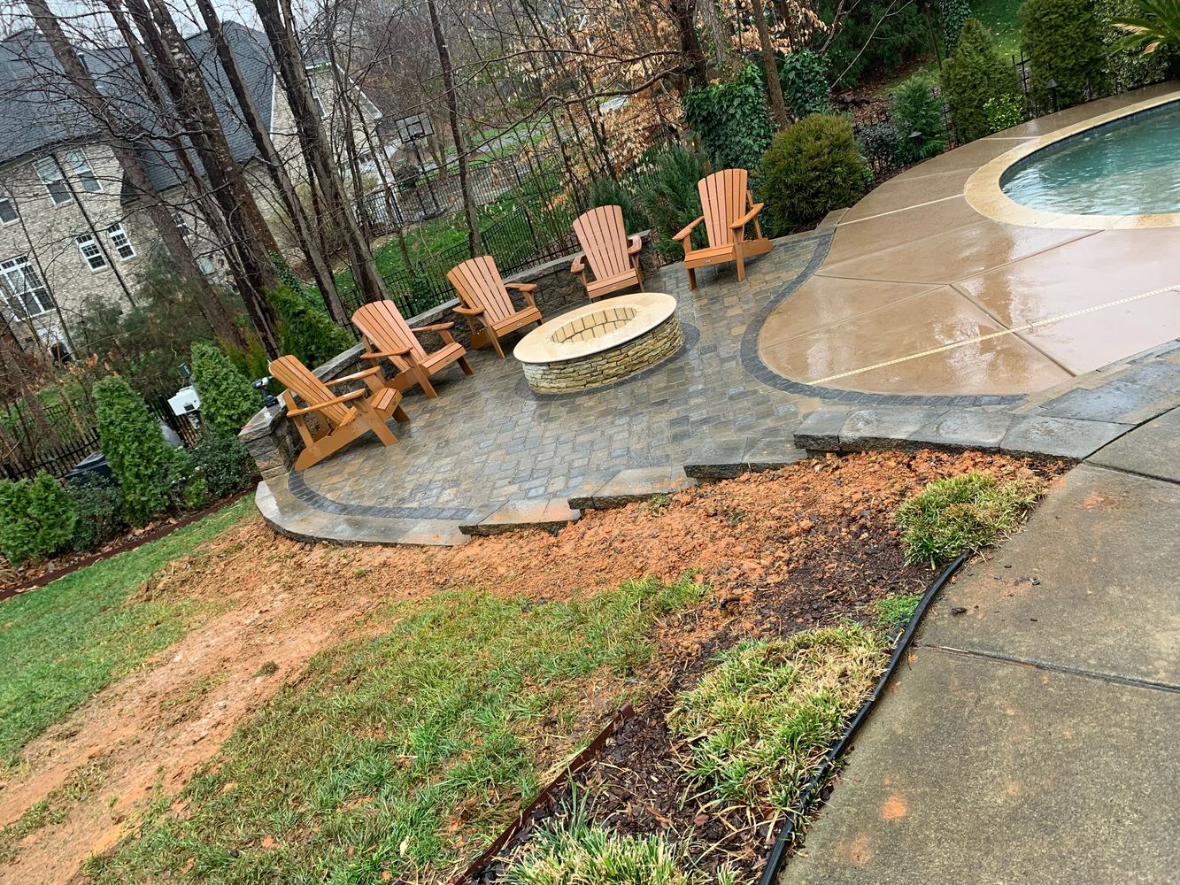 Backyard patio with wooden chairs around a stone fire pit beside a pool on a rainy day.