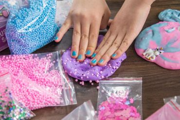 Hands playing with purple slime mixed with foam beads on a table with craft supplies.