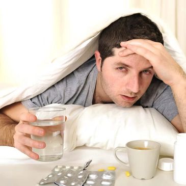 Man under blanket holding water, looking sick with medicines and coffee nearby.