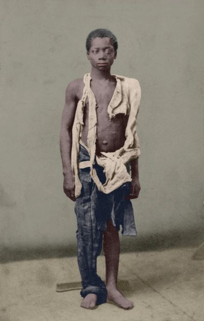 Portrait of a barefoot boy in tattered clothes against a plain backdrop.