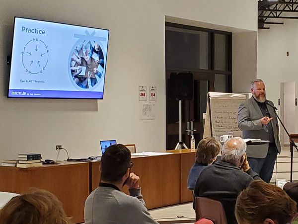 A man in a gray blazer speaks to an audience in a modern conference room.