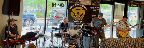 A four-member band performs live in a cozy indoor venue with large windows.