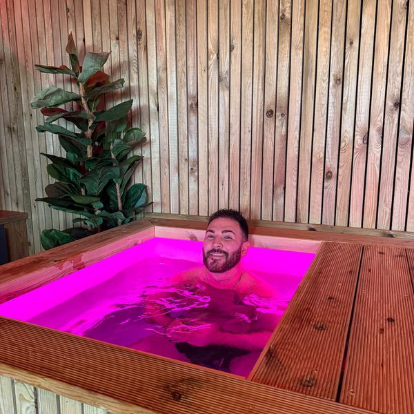 Man relaxing in a hot tub with vibrant pink lighting and wooden surroundings, practicing pranayama breathwork and cold water therapy