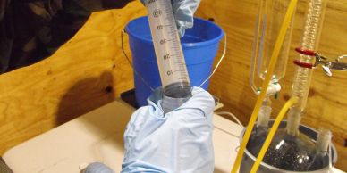 Gloved hand holding a graduated cylinder in a lab setting.