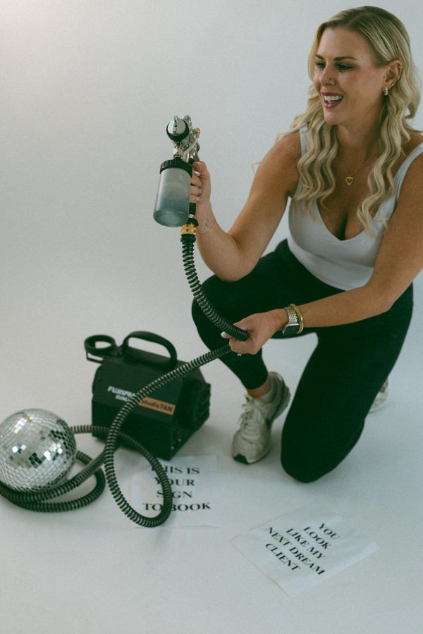 Woman holding a spray tanning gun with equipment and motivational notes on the floor.
