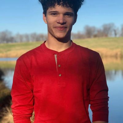 Young man in a red shirt smiling outdoors by a lake.