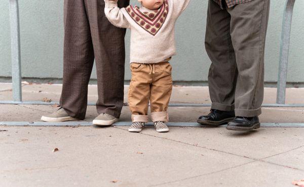 A toddler holding hands with two adults, learning to walk outdoors.