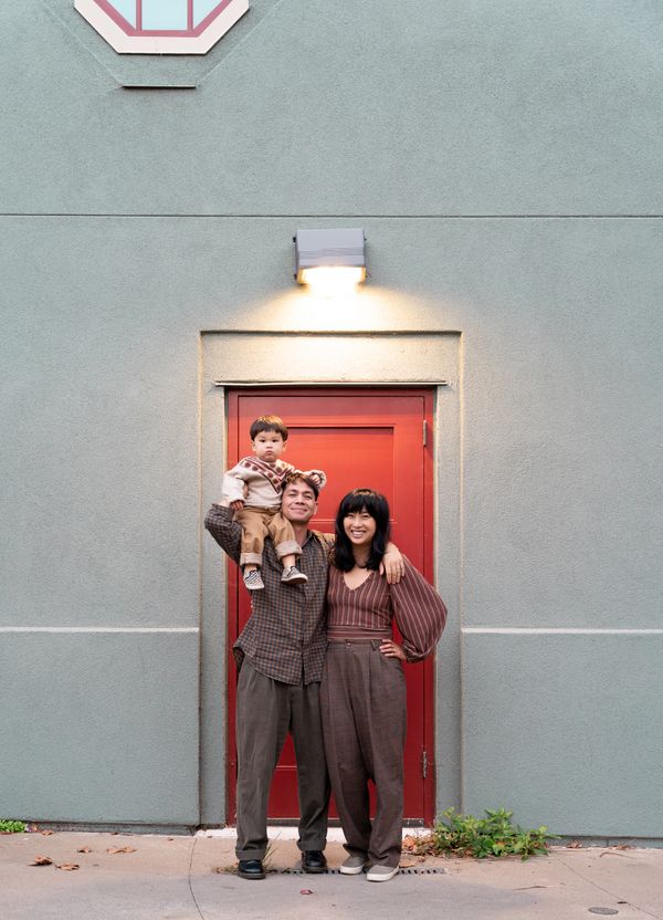 Happy family of three posing in front of a red door on a gray wall.