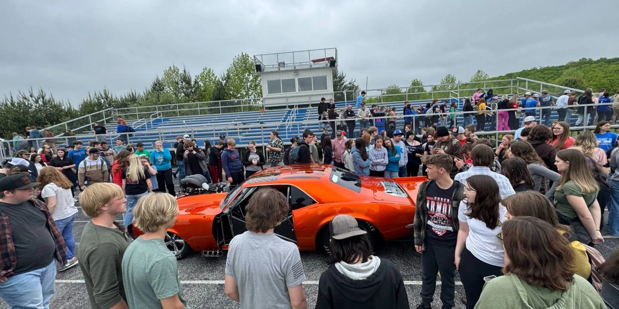Crowd gathered around a bright orange car at an outdoor event.