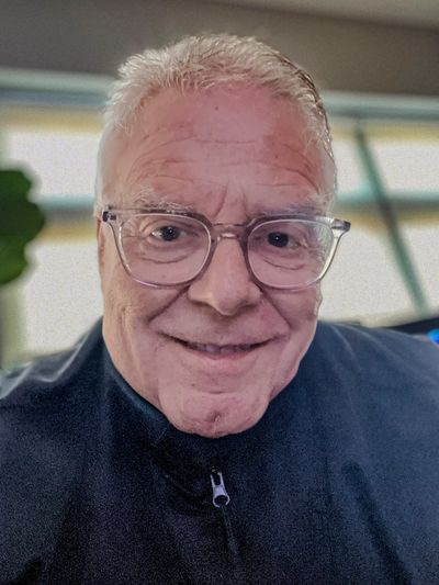 Smiling elderly man with glasses and gray hair in a dark jacket.